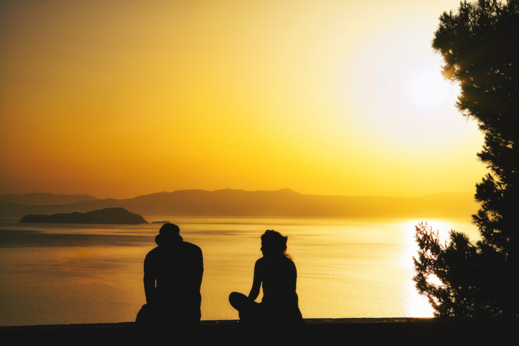 Zwei sitzende Menschen als Silhouette vor der Küste Kretas bei Chania zum Sonnenuntergang